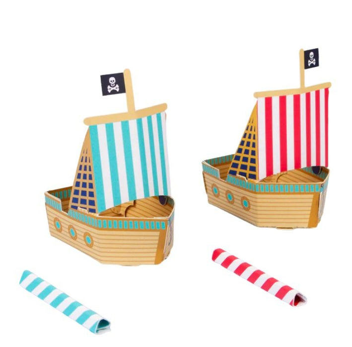 Image of two make your own pirate blow boats, one red and one blue. Featuring a blue and white stripe sail with jolly roger flag on the one boat and a red and white sail with jolly roger flag on the other. Image also shows a red stripey blow straw and a red stripey blow straw.