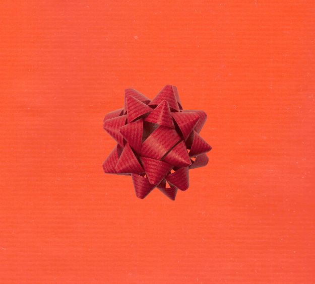 image of a square of wrapping paper, the paper is a solid orange kraft paper, in the centre of the gift wrap paper is a red paper gift wrapping bow