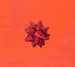 image of a square of wrapping paper, the paper is a solid orange kraft paper, in the centre of the gift wrap paper is a red paper gift wrapping bow