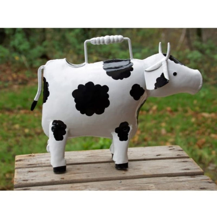 This lovely handcrafted, metal Cow Watering Can is painted white with black spots like a Friesian Cow. It has a metal carrying handle. It is made from painted iron sheet.  It is 21cm high, 35cm long, and 12cm wide. It is standing on a table in a garden.